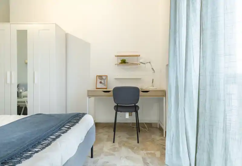 Camera da letto luminosa e minimal con letto ben rifatto, una scrivania compatta e un armadio; luce naturale diffusa e accenti blu creano un’atmosfera rilassante.
