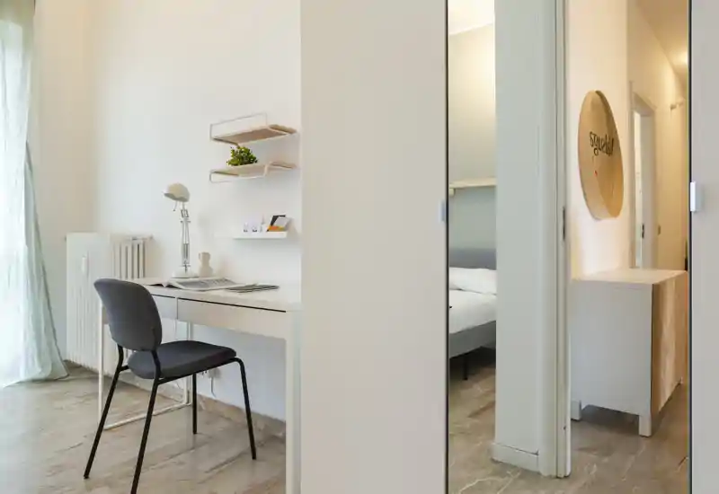 Bright, modern bedroom view seen through an open doorway: a portion of the bed is visible alongside a compact desk area with chair and shelving, creating a functional and minimal space.