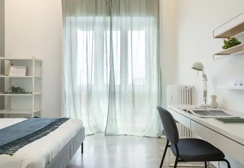 Bright, minimalist bedroom with a neatly made bed, soft teal throw and a desk by the window letting in abundant natural light through sheer curtains.