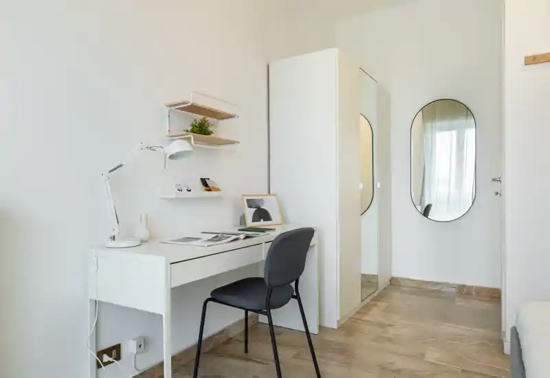 Bright, minimalist bedroom corner with a tidy white desk, chair, wardrobe with mirrored door and a glimpse of the bed at the frame edge; clean styling and natural light make it suitable for listing.
