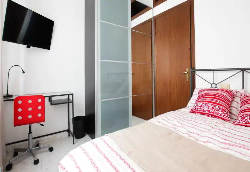 Bright, compact bedroom with a patterned bed, decorative cushions and a small desk with a red chair; wardrobe with mirrored panels and wall-mounted TV complete the practical layout.