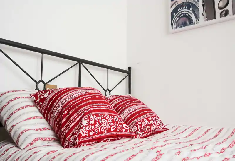 Bright, neatly styled bedroom with a decorative metal headboard and red patterned pillows on a white-and-red striped bedcover — cozy and well-lit for rental listing photos.