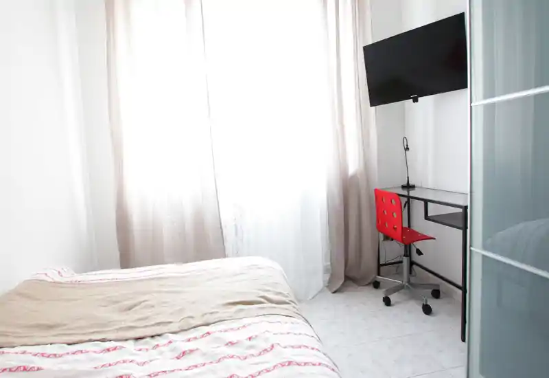 Bright, minimal bedroom with a neatly made bed, large window with sheer curtains, and a small desk with a red chair and wall-mounted TV.