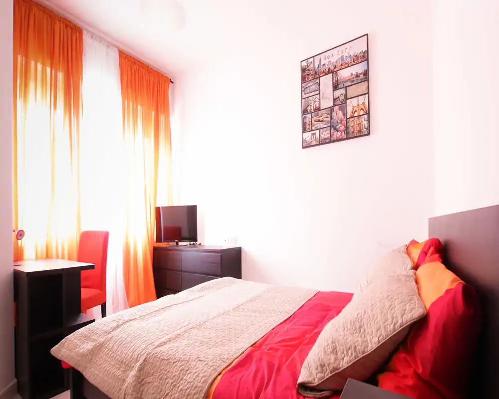 Bright, cozy bedroom with a double bed dressed in warm bedding, a dresser with TV and a small desk by a large window with orange curtains.