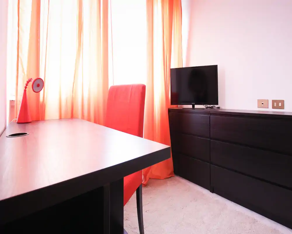 Bright, modern living area corner with a dark wood dresser, flat-screen TV and a desk with a red chair and lamp in front of sunlit orange curtains.