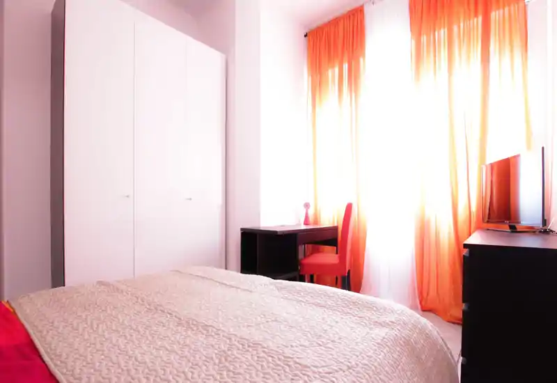 Bright, cozy bedroom with a large visible bed in the foreground, orange curtains, a desk and TV — ideal for a sunny rental listing.