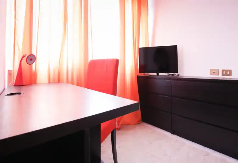 Bright, modern living area corner with a dark wood dresser, flat-screen TV and a desk with a red chair and lamp in front of sunlit orange curtains.