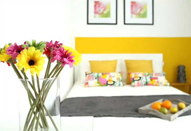 Bright, styled bedroom with a white bed, colorful cushions and a cheerful yellow headboard accent. A vase with vibrant flowers in the foreground adds a welcoming, photographed-for-listing feel.