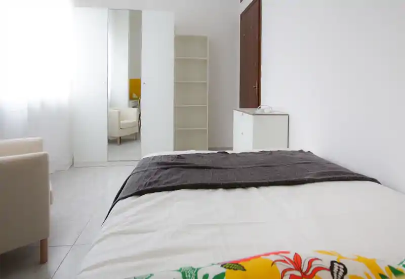 Bright minimalist bedroom with a large bed in the foreground, white furniture, and a mirrored wardrobe reflecting a cozy armchair.