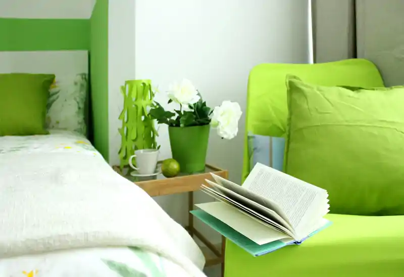 Coin chambre lumineux et soigné avec lit visible, accents verts, table de chevet avec plante et fauteuil confortable avec un livre ouvert—parfait pour un séjour reposant.