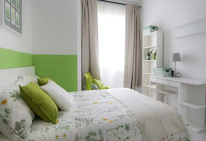 Chambre lumineuse et bien agencée avec couette florale, coussins verts et un coin bureau propre près de la fenêtre. La lumière naturelle et la palette blanc-vert offrent une ambiance chaleureuse idéale pour une annonce.