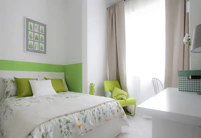 Chambre lumineuse et moderne avec grand lit en linge floral, coussins verts et un fauteuil vert près de la fenêtre — décoration épurée, fraîche et idéale pour une annonce.
