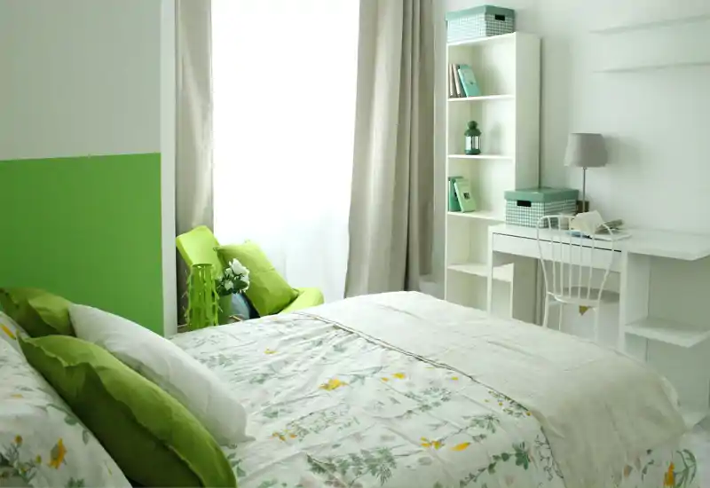Chambre lumineuse et cosy avec couette fleurie, coussins verts et un coin bureau rangé avec étagères — belle lumière naturelle venant de la fenêtre.