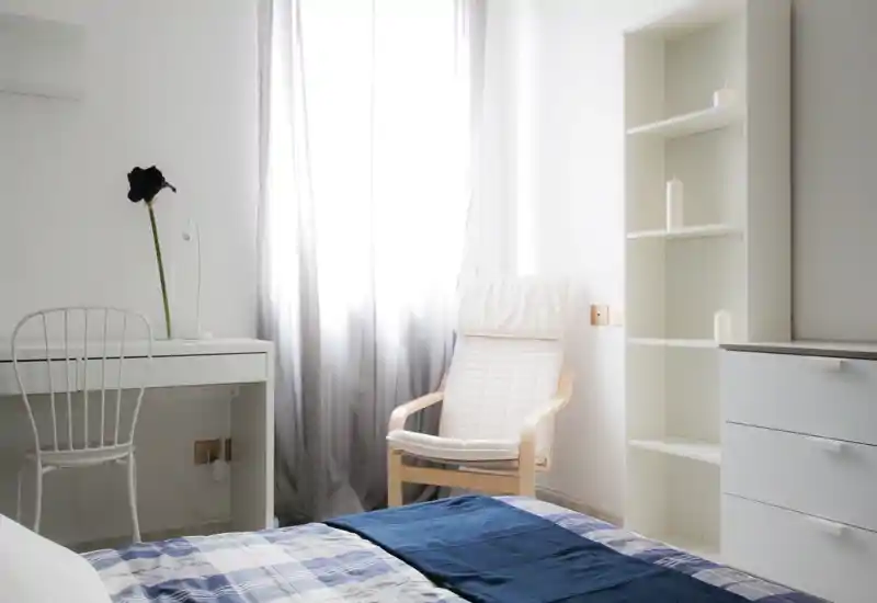 Dormitorio luminoso y minimalista con cama en primer plano, una butaca junto a la ventana y estantería blanca sencilla — ideal para una estancia tranquila y aireada.