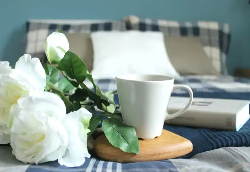 Bright, styled bedroom detail showing a cozy bed with pillows, a cup on a wooden tray and decorative white roses — ideal for a calm morning scene.
