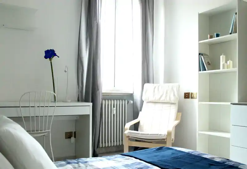 Bright, minimalist bedroom with a neatly made bed in the foreground, a comfortable armchair by the window and a simple desk with a single flower — airy, modern and inviting.