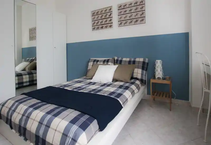 Bright, tidy bedroom with a double bed dressed in plaid linens, decorative cushions and a bedside table. Neutral walls with a blue accent panel and mirrored wardrobe create a sense of space.