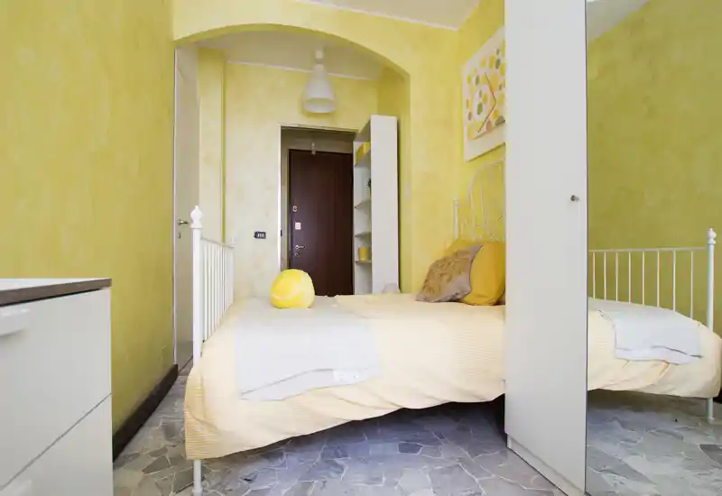 Camera singola luminosa e accogliente con pareti gialle, struttura letto in metallo bianco e biancheria morbida. Decorazioni e mensole vicino all'ingresso rendono lo spazio funzionale e soleggiato.