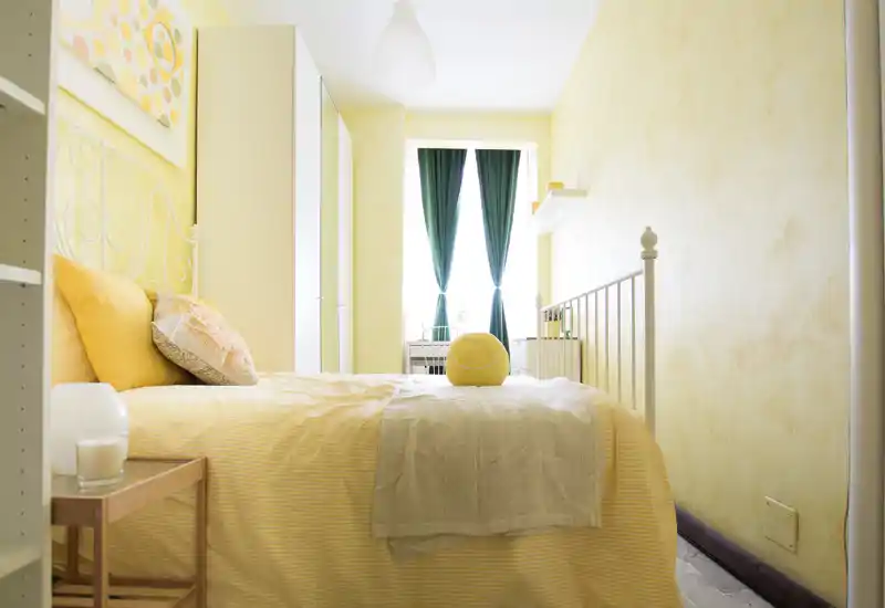 Camera da letto luminosa e accogliente con letto ben rifatto, tonalità calde gialle e tende verdi che lasciano entrare luce naturale — perfetta per un annuncio.