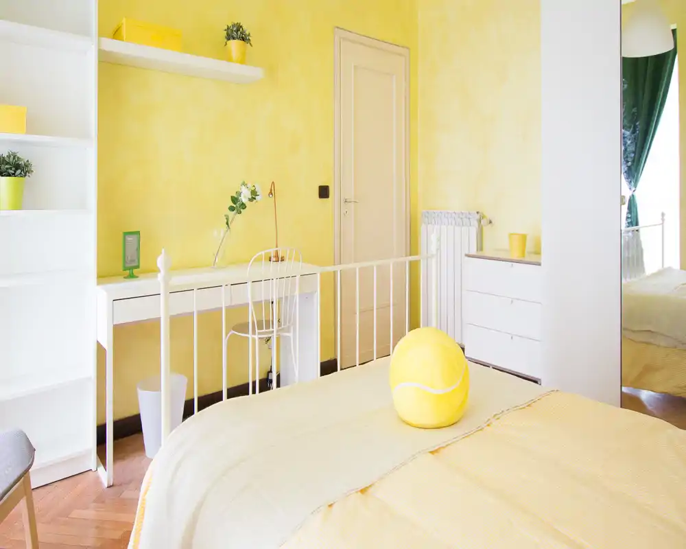 Bright, sunny bedroom with warm yellow walls and coordinated bedding. A neat metal bed frame, desk and shelving create a cheerful, functional space ideal for short-term rentals.