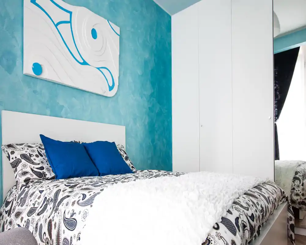 Dormitorio luminoso y cuidado con ropa de cama estampada, cojines azules y pared texturizada turquesa; armario blanco grande y luz natural que aportan sensación moderna y despejada.