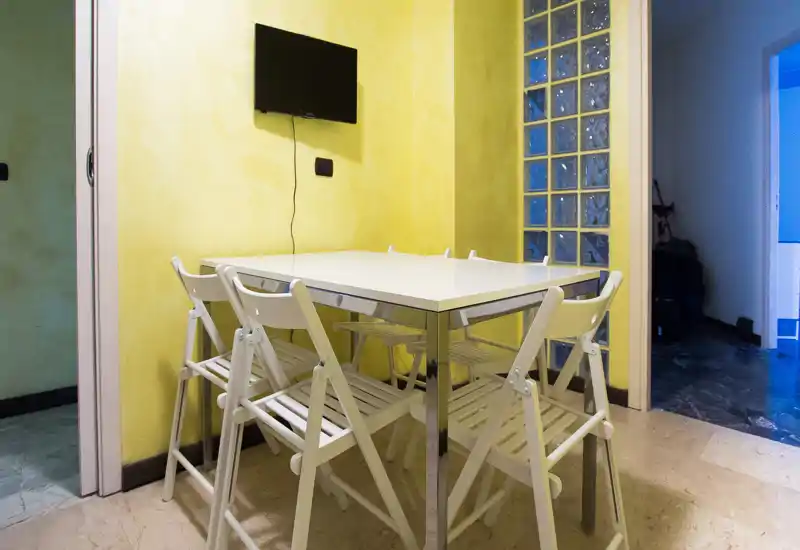 Piccola zona pranzo con tavolo quadrato bianco e sedie pieghevoli davanti a una parete gialla. Spazio pratico e luminoso adatto per pasti informali.