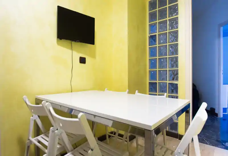 Zona pranzo compatta con tavolo bianco lucido e sedie pieghevoli, pareti giallo chiaro e TV a parete — pratica e ben illuminata per i pasti.