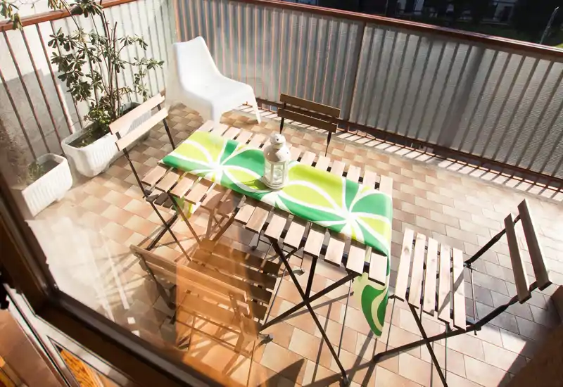 Balcone soleggiato con tavolo e sedie pieghevoli in legno, runner verde decorativo e vasi con piante — ideale per colazione o relax all’aperto.