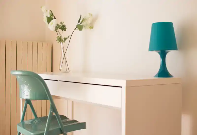 Angolo scrivania luminoso e minimal con un tavolo bianco, sedia pieghevole turchese e lampada coordinata. Un vaso di vetro con fiori bianchi aggiunge un tocco decorativo, perfetto per mostrare un piccolo spazio di lavoro.