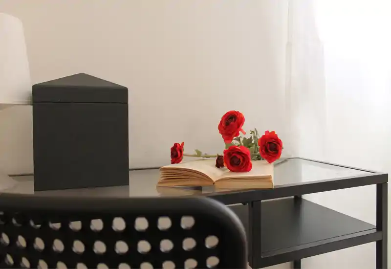 Angolo luminoso e minimal con scrivania in vetro, fiori rossi decorativi e un libro aperto che creano un angolo di lavoro tranquillo e accogliente.