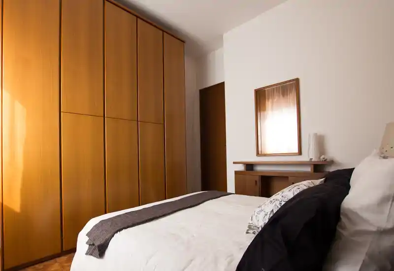 Camera da letto luminosa e accogliente con un letto matrimoniale ben rifatto, un ampio armadio in legno e un piccolo comò con specchio; la luce naturale da sinistra crea un'atmosfera calda.