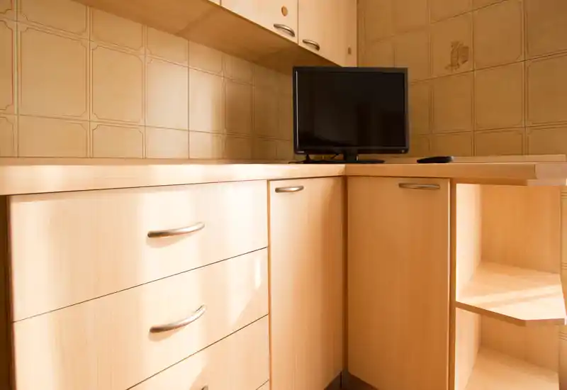 Angolo cucina compatto con mobili in legno chiaro, cassetti e una piccola TV sul piano di lavoro; piastrelle calde e superfici ordinate creano uno spazio funzionale e accogliente.