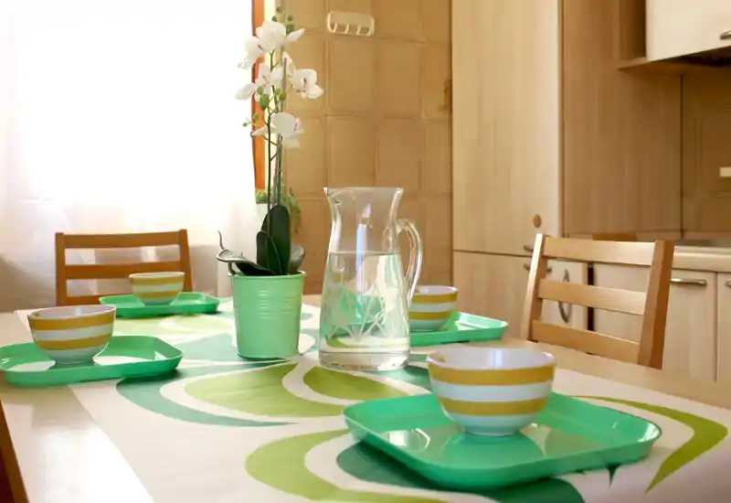 Zona pranzo luminosa e curata: vassoi verdi e ciotole a righe su una tovaglia decorata, brocca d'acqua in vetro e un'orchidea in vaso per un'atmosfera fresca e accogliente.