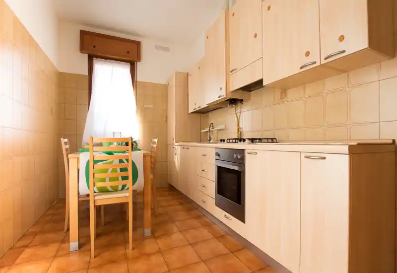 Cucina compatta e luminosa con mobili in legno chiaro, forno incorporato e un piccolo tavolo da pranzo vicino alla finestra — piastrelle calde e luce naturale creano un'atmosfera accogliente.