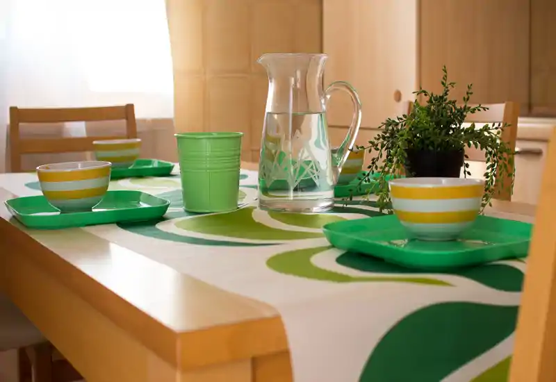 Tavolo da pranzo luminoso e ben arredato con stoviglie a righe verdi, una caraffa d'acqua in vetro e una piccola pianta — perfetto per pasti informali in un angolo accogliente.