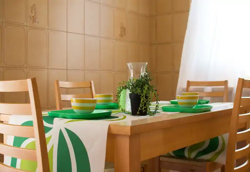 Zona pranzo luminosa e curata con tavolo in legno per quattro persone, stoviglie con accenti verdi e una piccola pianta al centro—perfetta per pasti informali.