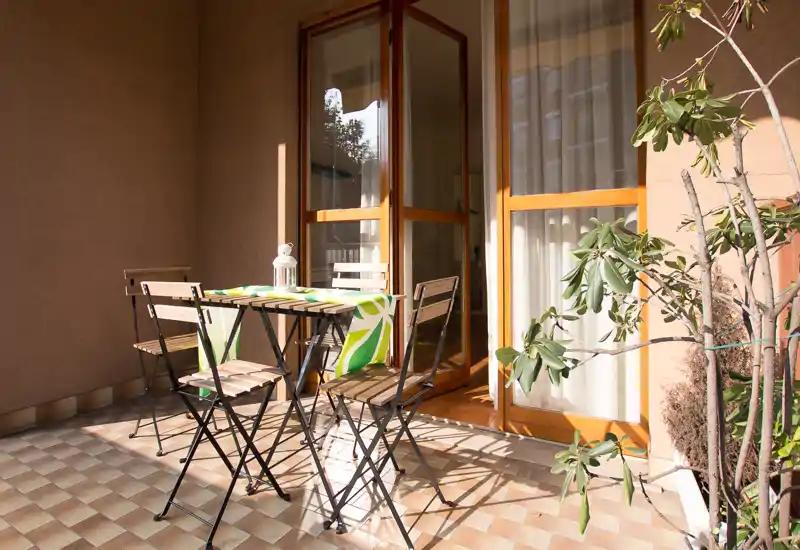 Balcone soleggiato arredato con un piccolo tavolo pieghevole e quattro sedie in legno, runner decorativo e piante in vaso — ideale per il caffè del mattino.