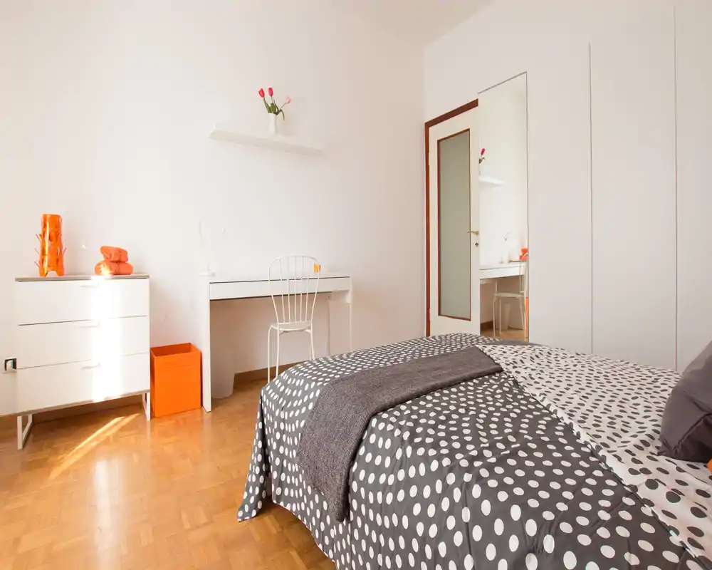 Camera da letto moderna e luminosa con letto matrimoniale a fantasia, scrivania e comò bianchi e pavimento in parquet; dettagli semplici donano un'atmosfera fresca e accogliente.