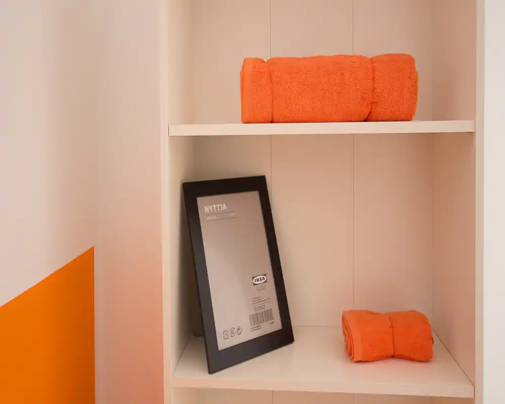 Primo piano di una mensola bianca con asciugamani arancioni ripiegati e una cornice con etichetta IKEA appoggiata. L'immagine mostra dettagli di scaffalatura e cromia, non una stanza completa.