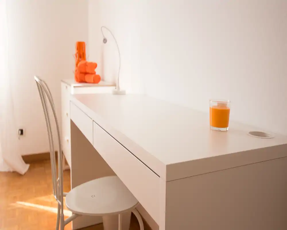 Angolo living/ufficio luminoso e minimale con una scrivania bianca, sedia in metallo e accenti arancio (asciugamani e candela). La composizione evidenzia uno spazio di lavoro pulito e arioso, illuminato con luce naturale.