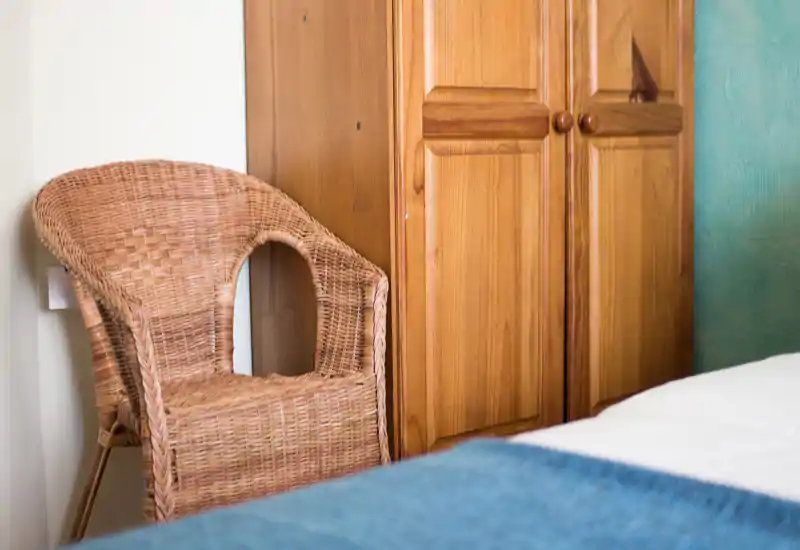 Angolo della camera accogliente con vista parziale del letto, una sedia in vimini e un armadio in legno, illuminato da una luce naturale morbida.