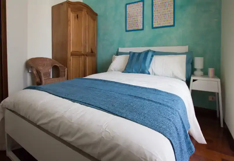 Camera da letto luminosa e ben curata con letto matrimoniale in biancheria bianca e blu, armadio in legno e comodino — arredo moderno e rilassante, ideale per il riposo.