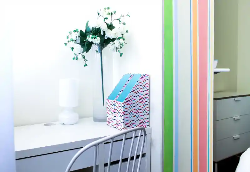 Angolo di camera con scrivania bianca, lampada decorativa e vaso di fiori; luminoso e dallo stile minimal, adatto a un ambiente notte compatto.