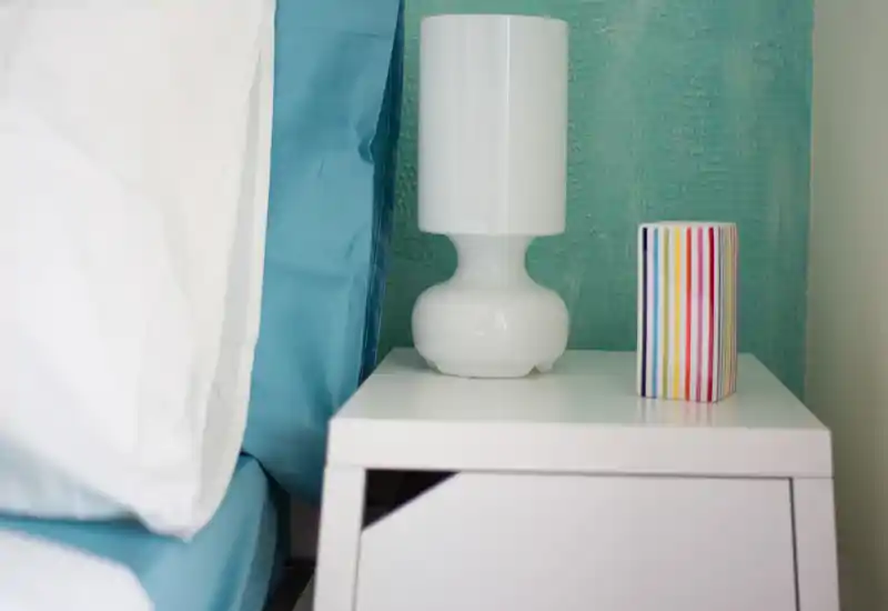 Dettaglio luminoso e minimale della camera: comodino bianco con lampada e un oggetto decorativo a righe, accanto a cuscino e lenzuola blu.