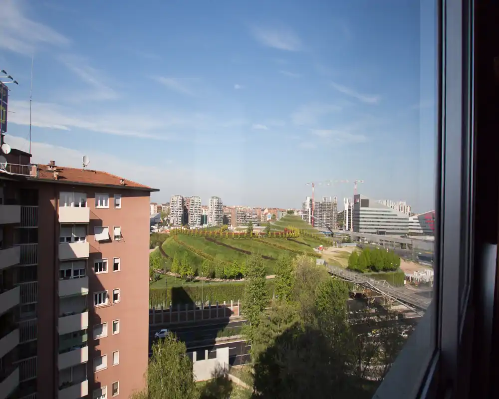 Bright cityscape view from a window showcasing a landscaped green hill, surrounding residential blocks and modern buildings — ideal to highlight the apartment's urban view.