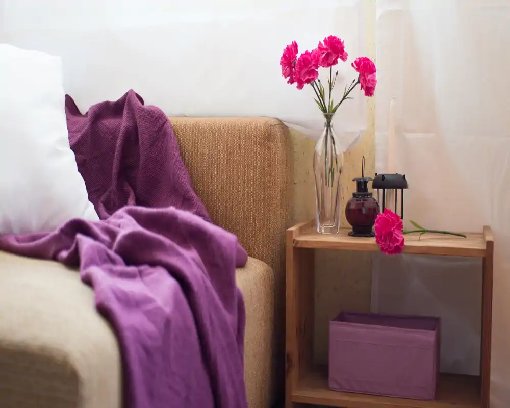 Cozy corner of a living room with a soft sofa draped in a purple throw, a decorative cushion, and a small wooden side table topped with fresh flowers and candles — bright, styled and inviting.