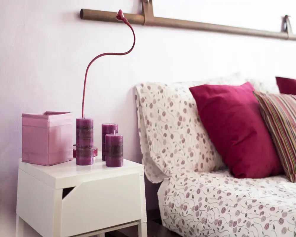 Bright, cozy bedroom corner with patterned bedding, plush magenta cushions and a white bedside table decorated with purple candles — ideal for a warm, stylish rental listing.