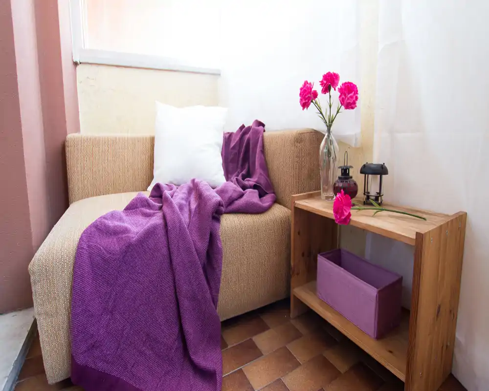 Cozy corner with a beige upholstered sofa chair, accented by a purple throw and white cushion, plus a small wooden side shelf with decorative flowers — a bright, inviting living nook.
