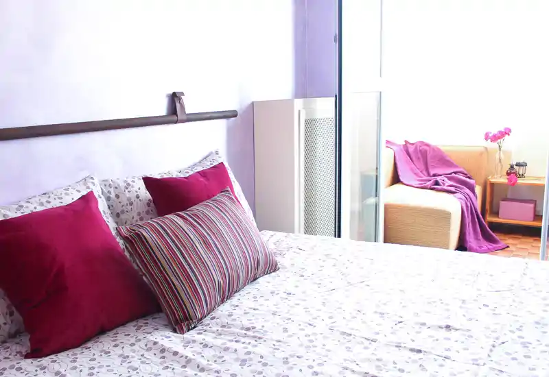 Camera da letto luminosa e accogliente con copriletto a fiori, cuscini magenta e un cuscino a righe; luce naturale e un piccolo angolo salotto vicino alla finestra.