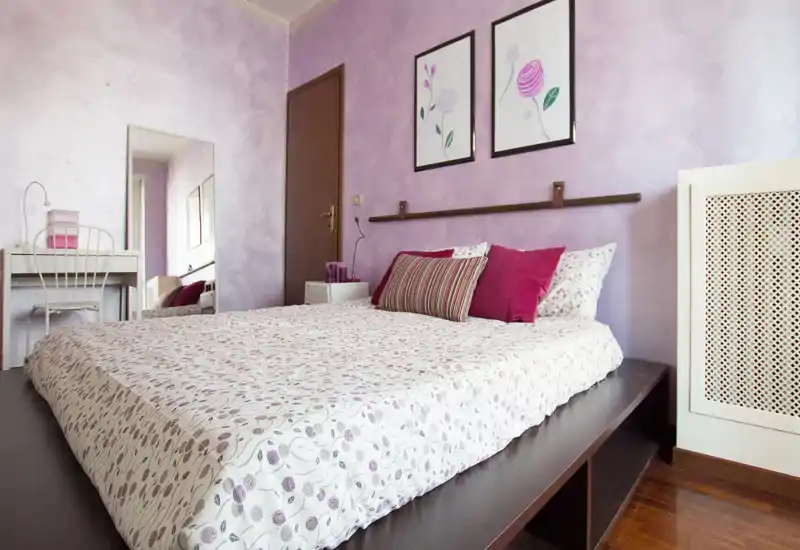 Camera da letto luminosa e femminile con letto su pedana, biancheria a fantasia e cuscini decorativi su pareti lilla. Il locale comprende una scrivania con specchio e pavimento in legno caldo, ambiente accogliente e ben allestito.